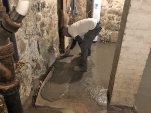 Person smoothing wet concrete on a basement floor with a trowel. The setting has exposed stone walls and pipes, creating a rustic, under-construction feel.